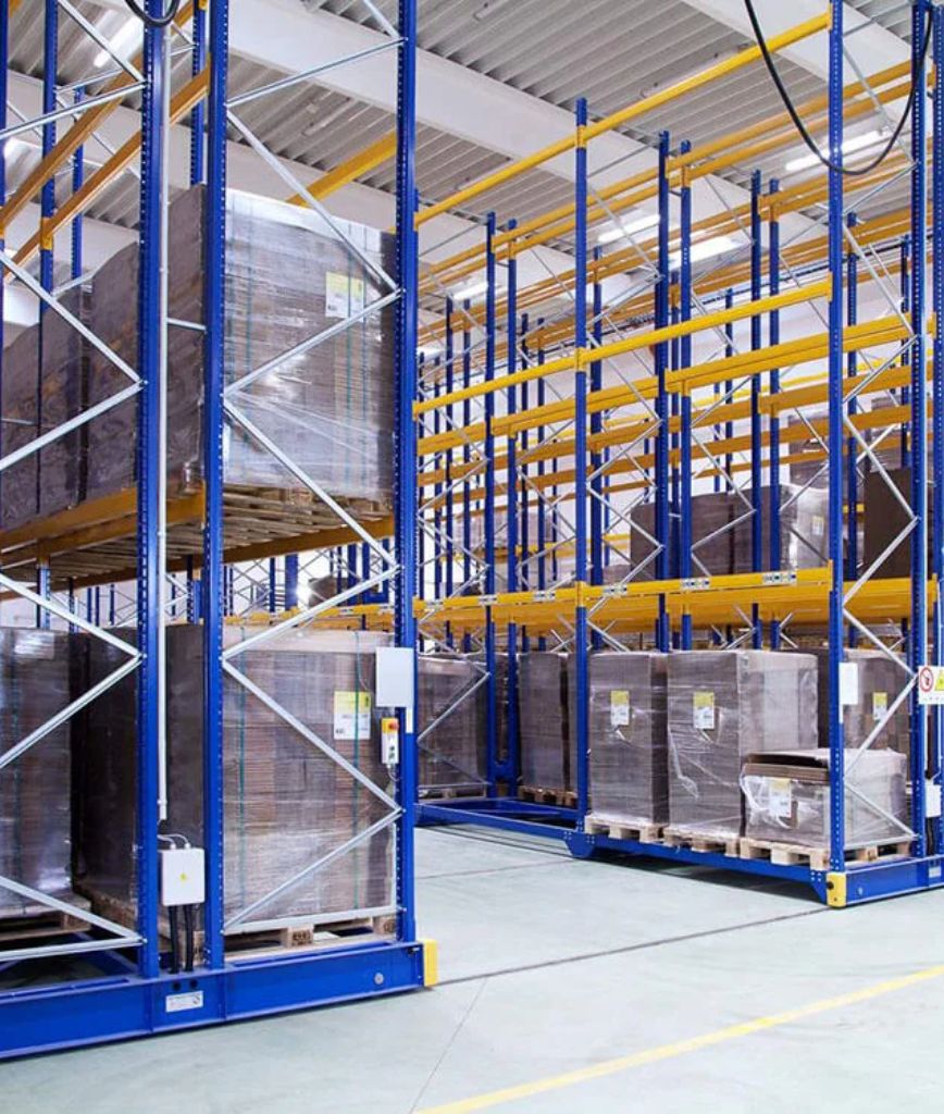 A spacious warehouse belonging to a pillow distributor, with tall blue and yellow storage racks holding large pallets wrapped in plastic, arranged neatly in organized rows under a high ceiling.