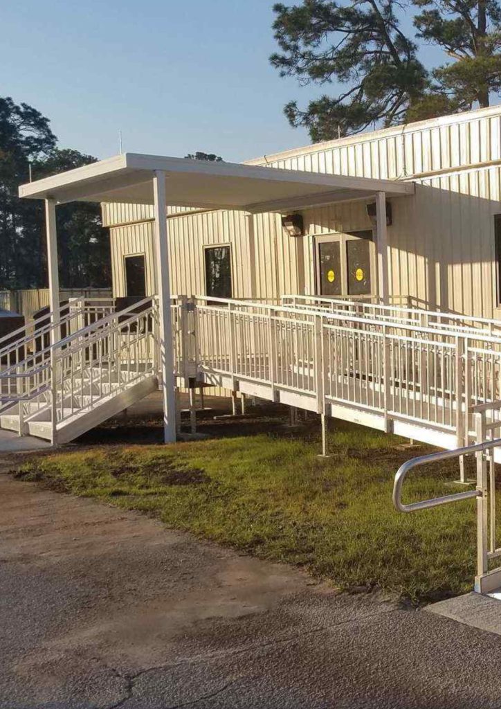 A beige prefabricated office building with two accessible ramps and white railings leads to a covered entrance. Yellow circles decorate the doors, with trees in the background—an example of fast construction solutions.