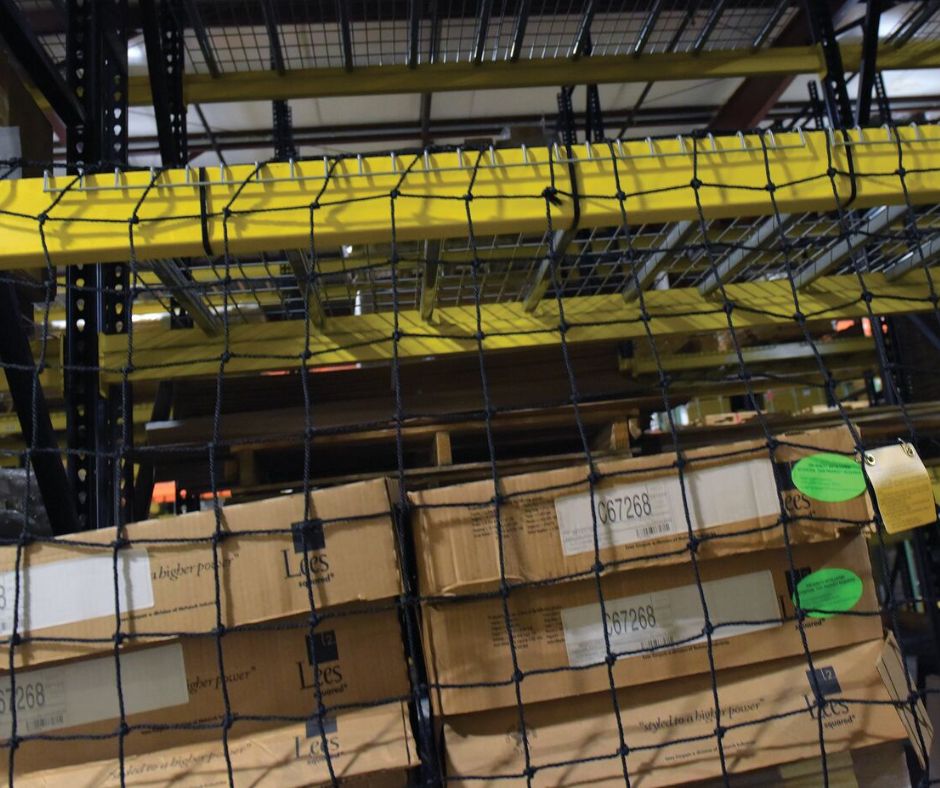Cardboard boxes are stacked on industrial shelves in a warehouse, secured by a black safety net Pallet Rack Backing. Yellow metal bars support the shelving above, and the boxes have labels and green tags attached.