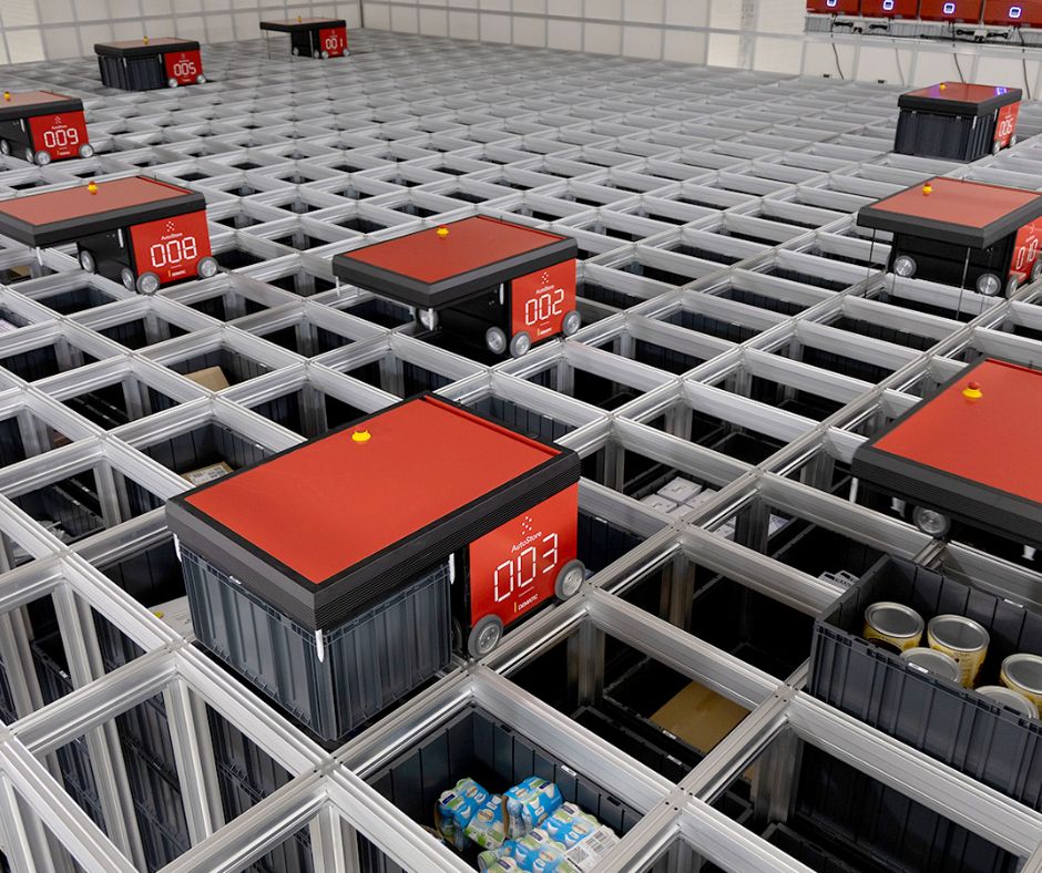Several red automated robots move along a grid of metal tracks, sorting and retrieving grocery items such as milk and canned goods in a smart warehouse design that features advanced warehouse automation solutions.