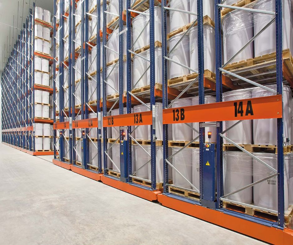 A large warehouse with tall, orange and blue metal racks stacked with palletized goods from a pillow distributor, wrapped in plastic, labeled with numbers and letters, and an empty aisle running alongside the shelves.