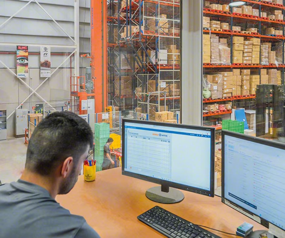 A man sits at a desk with two computer monitors in an office overlooking a large warehouse designed for CGMP warehouse compliance, filled with shelves stocked with boxes and various items.