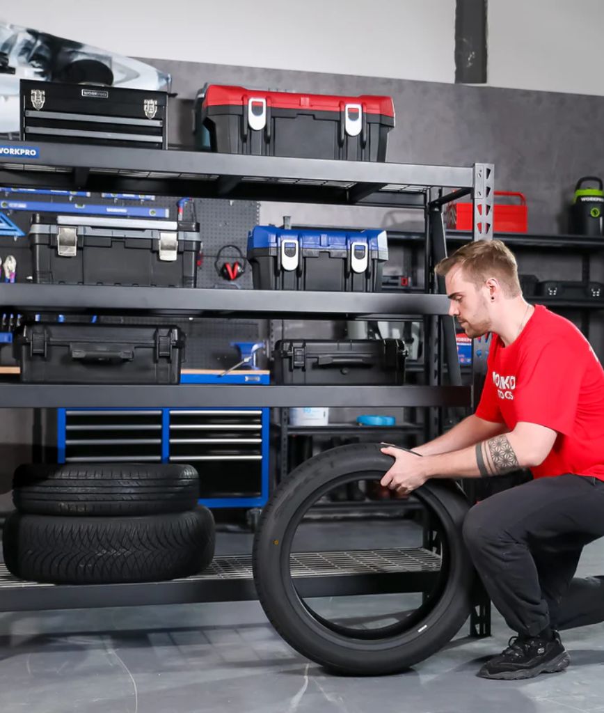 A man in a red shirt lifts a car tire in an automotive parts room design, with toolboxes and tires organized on metal shelves behind him in the garage workshop.
