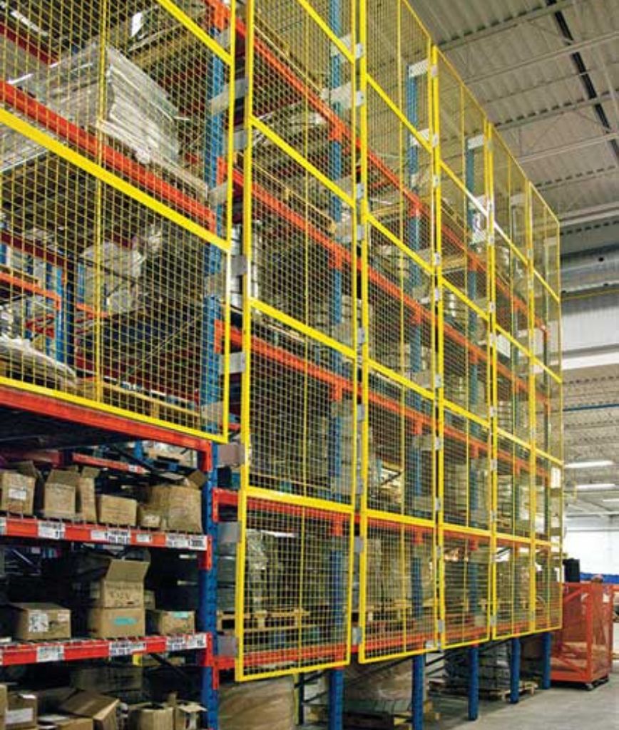 Tall warehouse storage racks filled with various boxes and items are secured with yellow pallet rack backing mesh panels, inside a brightly lit industrial facility with a high ceiling.