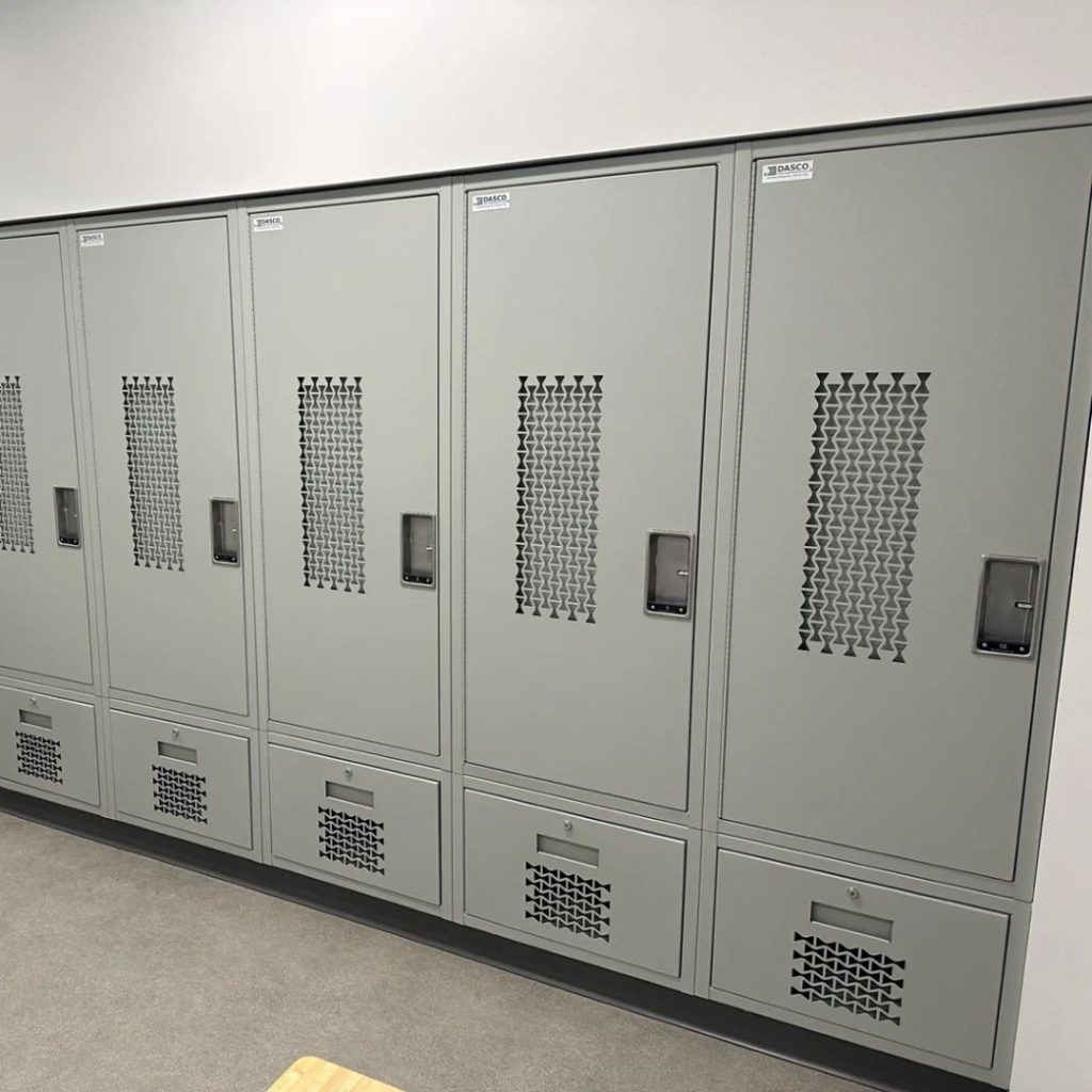 A row of six gray metal lockers with ventilation grilles, name labels, and small storage compartments at the bottom, situated in a clean, well-lit room with a beige floor.