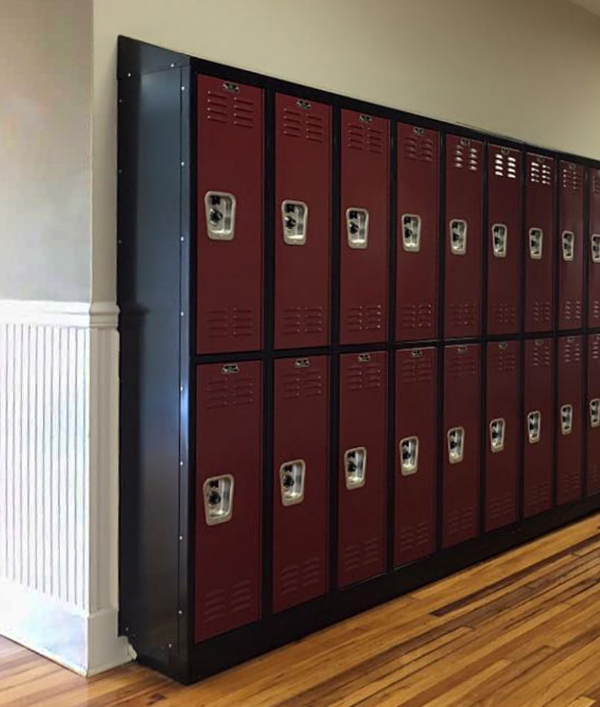 A row of maroon school lockers with silver handles is lined up against a beige wall with white paneling. The lockers are above a polished wooden floor in a hallway.