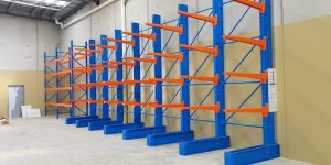 Blue and orange industrial cantilever racks are installed along a beige wall in a spacious warehouse with a concrete floor and high ceiling. The racks are empty and ready for storage use.