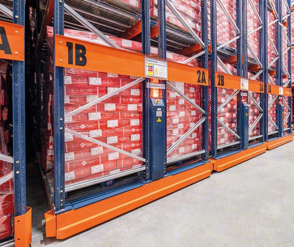 Warehouse shelving with orange and blue racks labeled 1B, 2A, and 2B holds stacks of red-wrapped products on pallets, organized in multiple rows on a concrete floor—ideal for implementing OSHA compliant warehouse design.