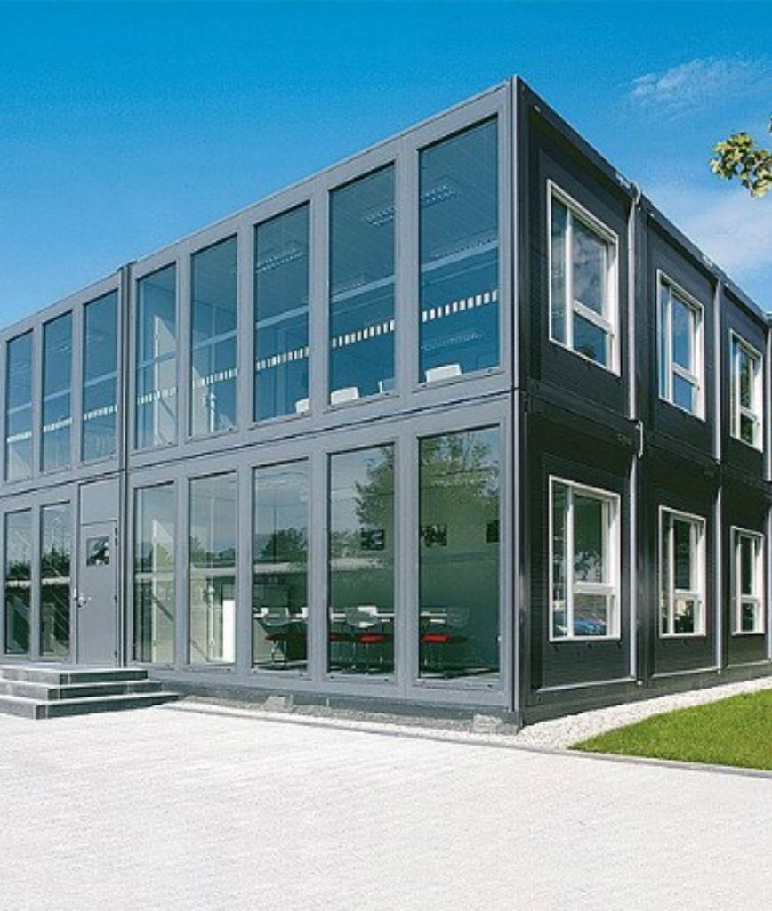 A modern two-story building made from stacked shipping containers, this sustainable prefabricated building features large glass windows and doors, set against a blue sky with a paved walkway and nearby greenery.