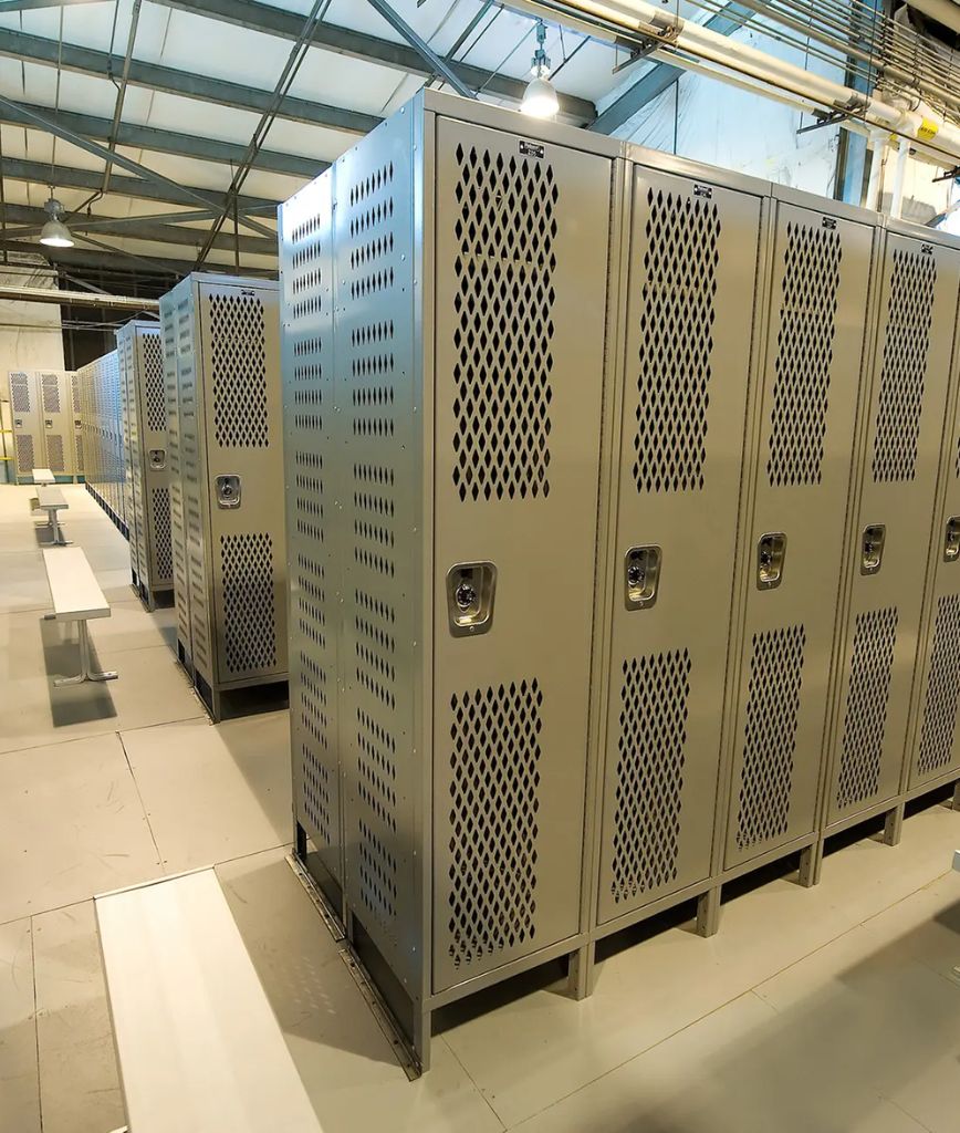 Rows of metal lockers with diamond-shaped ventilation holes in a brightly lit locker room, with white benches along the aisles and an exposed ceiling above.