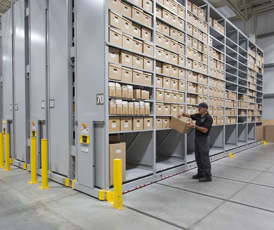 A worker in a uniform retrieves a cardboard box from a high-density mobile shelving system, showcasing warehouse capacity optimization in an organized warehouse with narrow aisle racking and yellow safety bollards lining the shelving units.