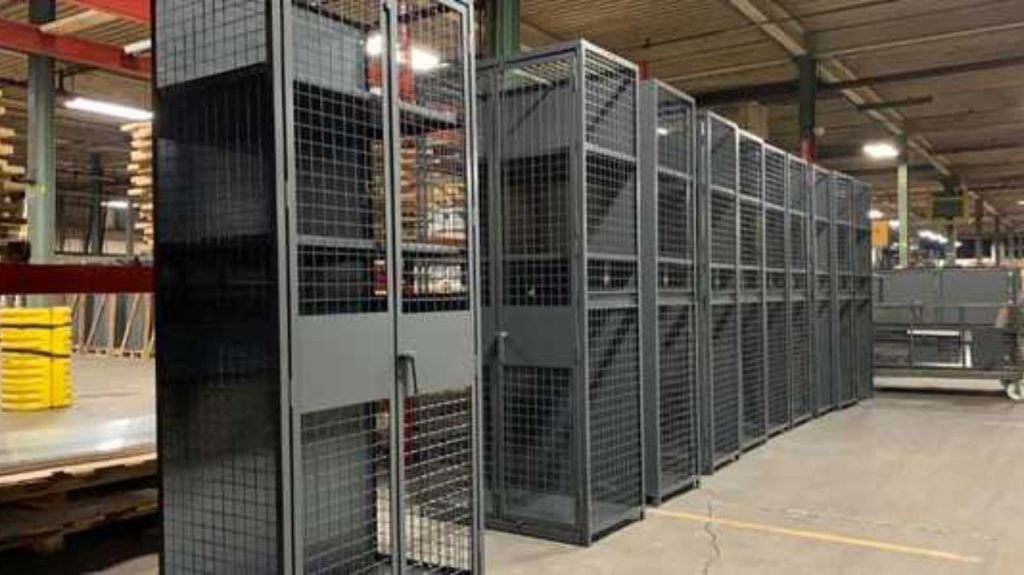 Rows of tall, black metal wire cages stand side by side in a large industrial warehouse with a high ceiling and exposed beams. The floor is concrete and various equipment and materials are visible in the background.
