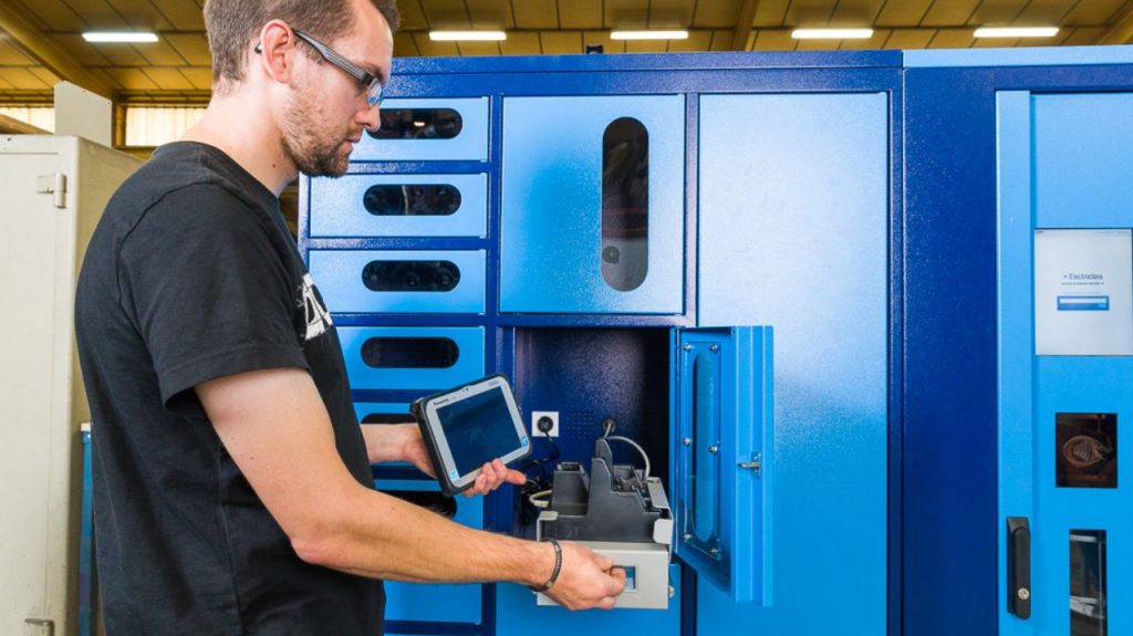 A person wearing safety glasses uses a touchscreen tablet while opening a blue industrial cabinet with multiple compartments in a workshop setting.