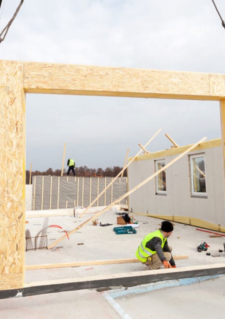 Two construction workers in safety vests assemble a building frame using a modular building process; one is on the ground floor and the other stands on a higher platform, surrounded by tools and wooden beams. The sky is cloudy.
