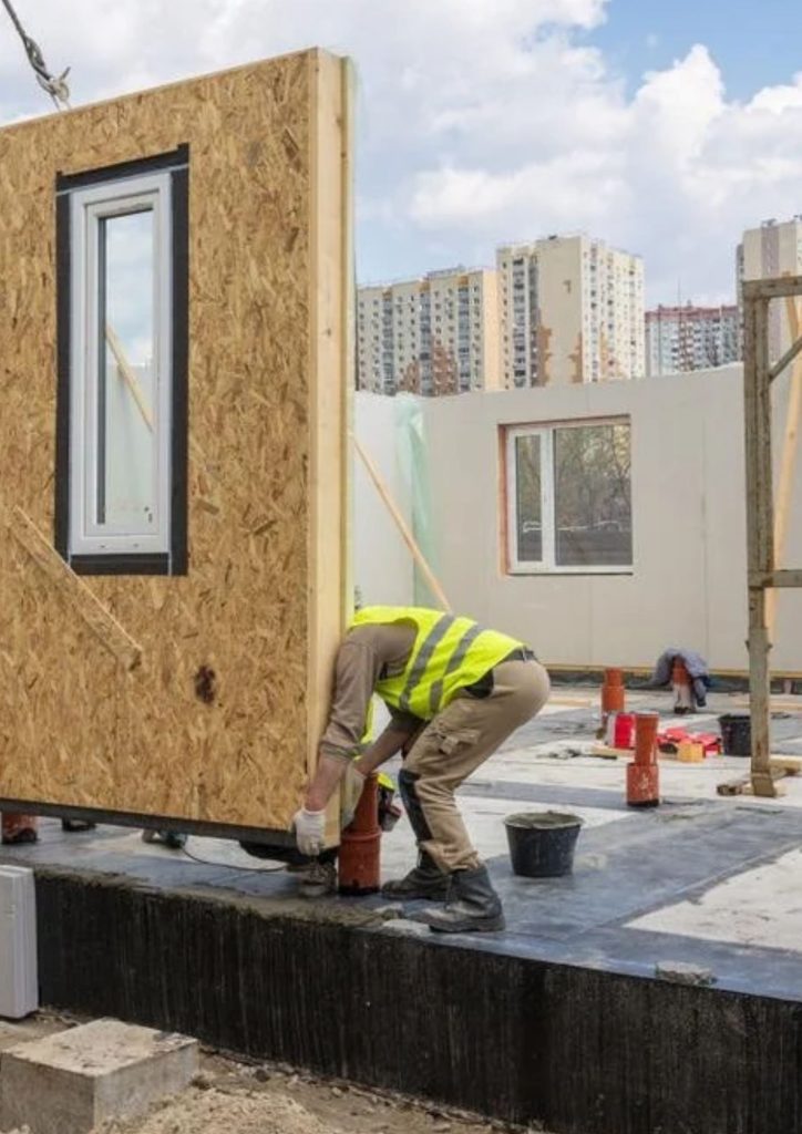 A construction worker in a yellow safety vest installs a prefabricated wall with a window on a building site, highlighting fast construction methods. Tall apartment buildings rise in the background beneath a partly cloudy sky.
