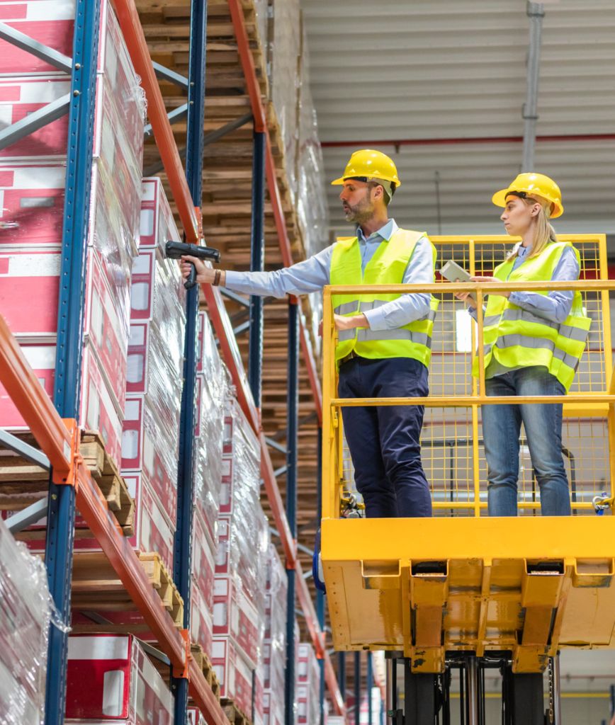 Two workers in yellow safety vests and hard hats stand on a warehouse lift. One scans boxes on high shelves using a barcode scanner, demonstrating smart warehouse design and efficient warehouse layout optimization.