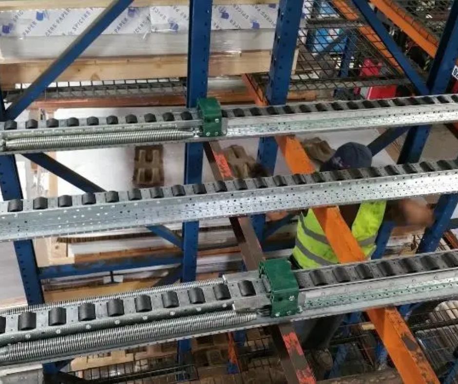 Warehouse shelving with metal roller tracks and orange beams, viewed from above, showcases a Pushback Pallet Racking System. A person in a yellow safety vest is partially visible, working among the shelves with pallets and materials stored nearby.