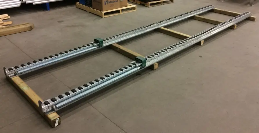 A metal roller track conveyor with inclined rails features two parallel rails and small black rollers, mounted on wooden beams and set on a concrete floor in an industrial setting.