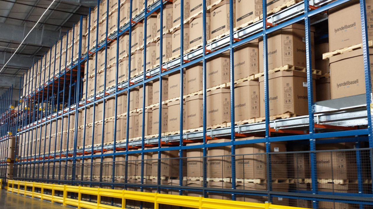Large warehouse features tall blue and orange metal racks in a narrow aisle racking layout, stacked with neatly organized brown cardboard boxes on wooden pallets. A yellow safety barrier runs along these high-density storage solutions.