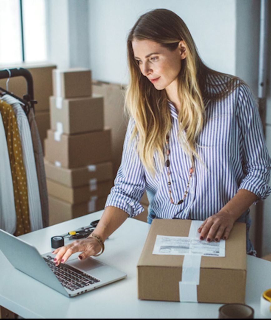 A woman stands at a desk using a laptop with one hand and holding a cardboard box with a shipping label in the other. She is surrounded by more boxes and clothing racks in a well-lit room.