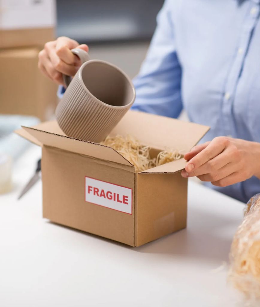 A person places a gray ceramic mug into a cardboard box labeled FRAGILE, with packing straw inside, preparing it for shipping or moving.