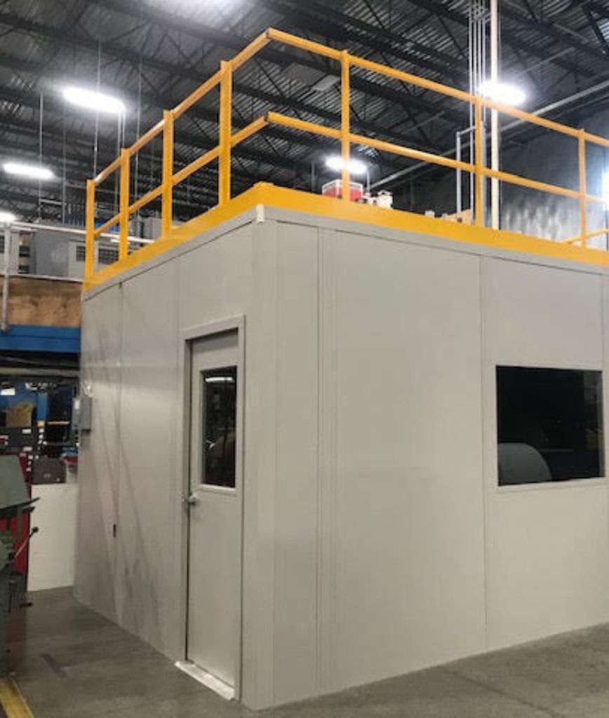 A white prefabricated office building with a door and window sits inside a warehouse, featuring a yellow-railed platform on its roof and industrial equipment in the background—showcasing the versatility of commercial modular buildings.