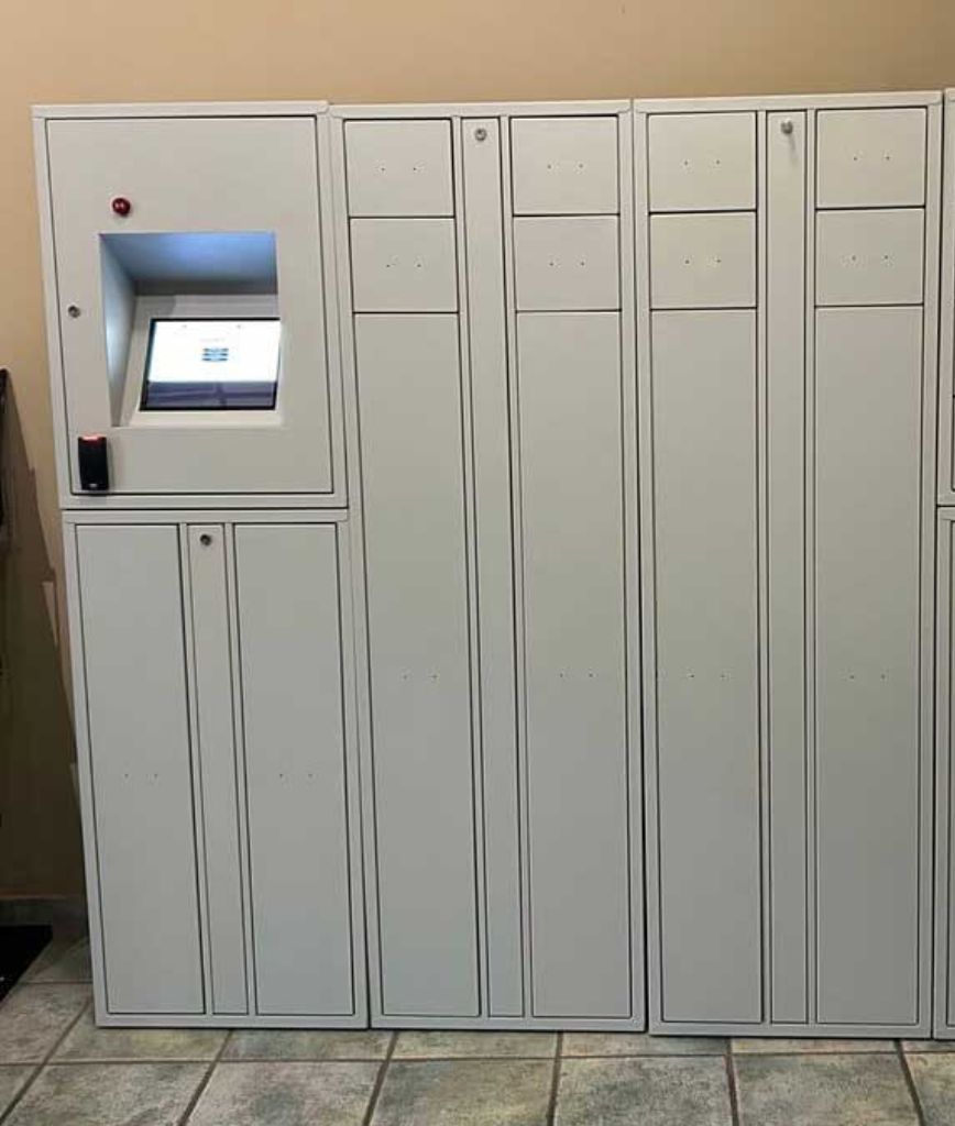 A set of white Secure Armory Solutions parcel locker units with a touchscreen interface on the upper left side, placed against a beige wall on a tiled floor.