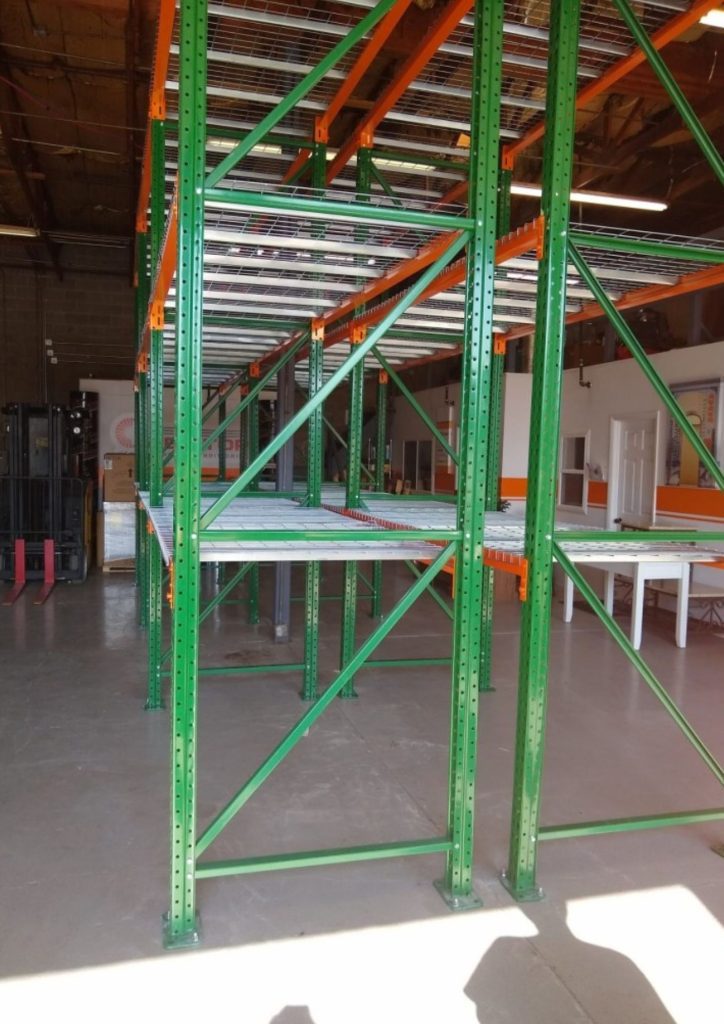 Empty green and orange Industrial Storage Racks Utah are set up inside a large building with concrete floors, exposed ceiling, and abundant natural light.