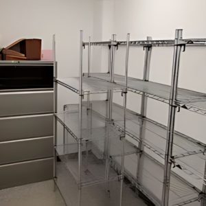 Adjustable wire shelving units and a gray filing cabinet stand in a small, white room. The shelves are empty, with just a few files and papers stacked on top of the cabinet.
