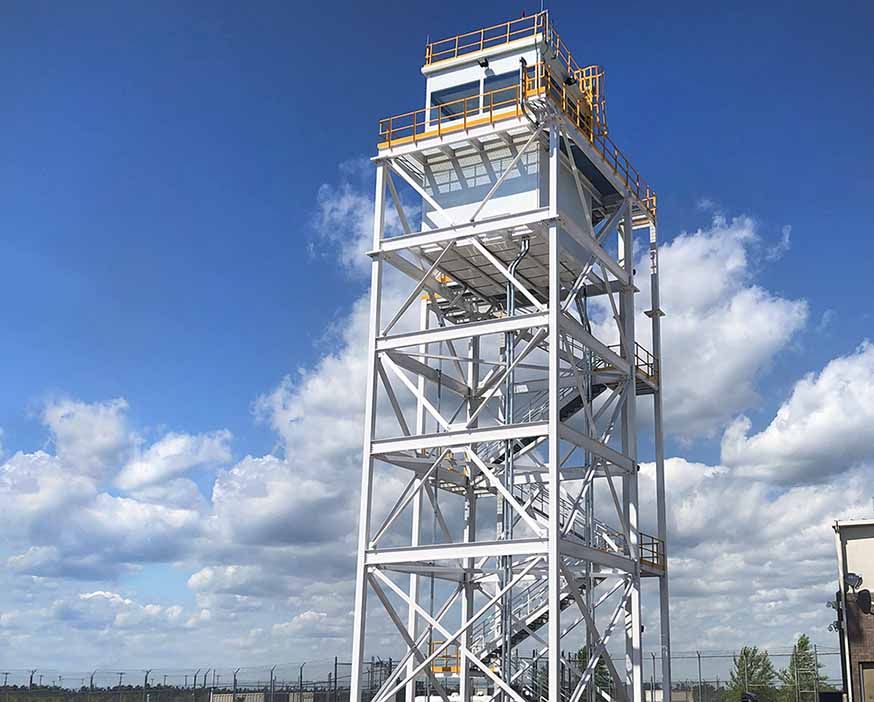 Galvanized steel lattice lookout tower with an enclosed cab and railed platform against a blue sky
