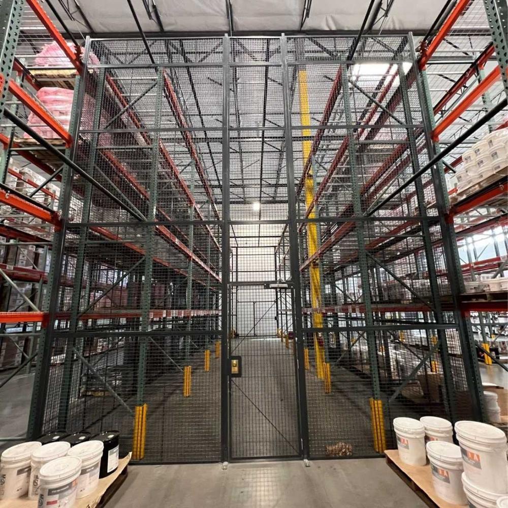 Tall wire mesh security enclosure spanning a pallet rack aisle with pails of coating stacked nearby