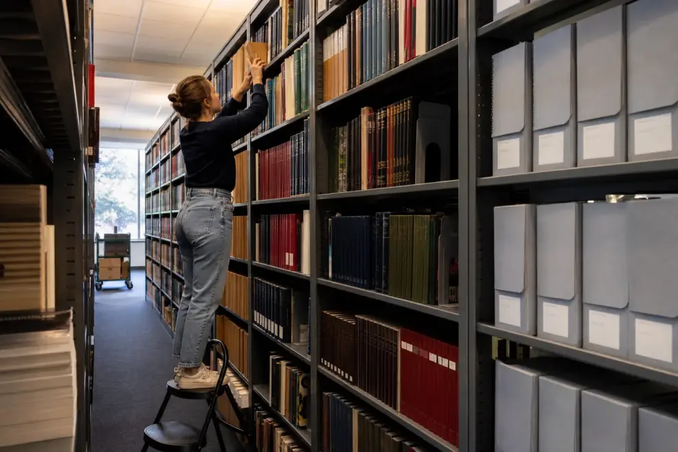 Worker on a step stool retrieving archival volumes from a tall steel shelving range