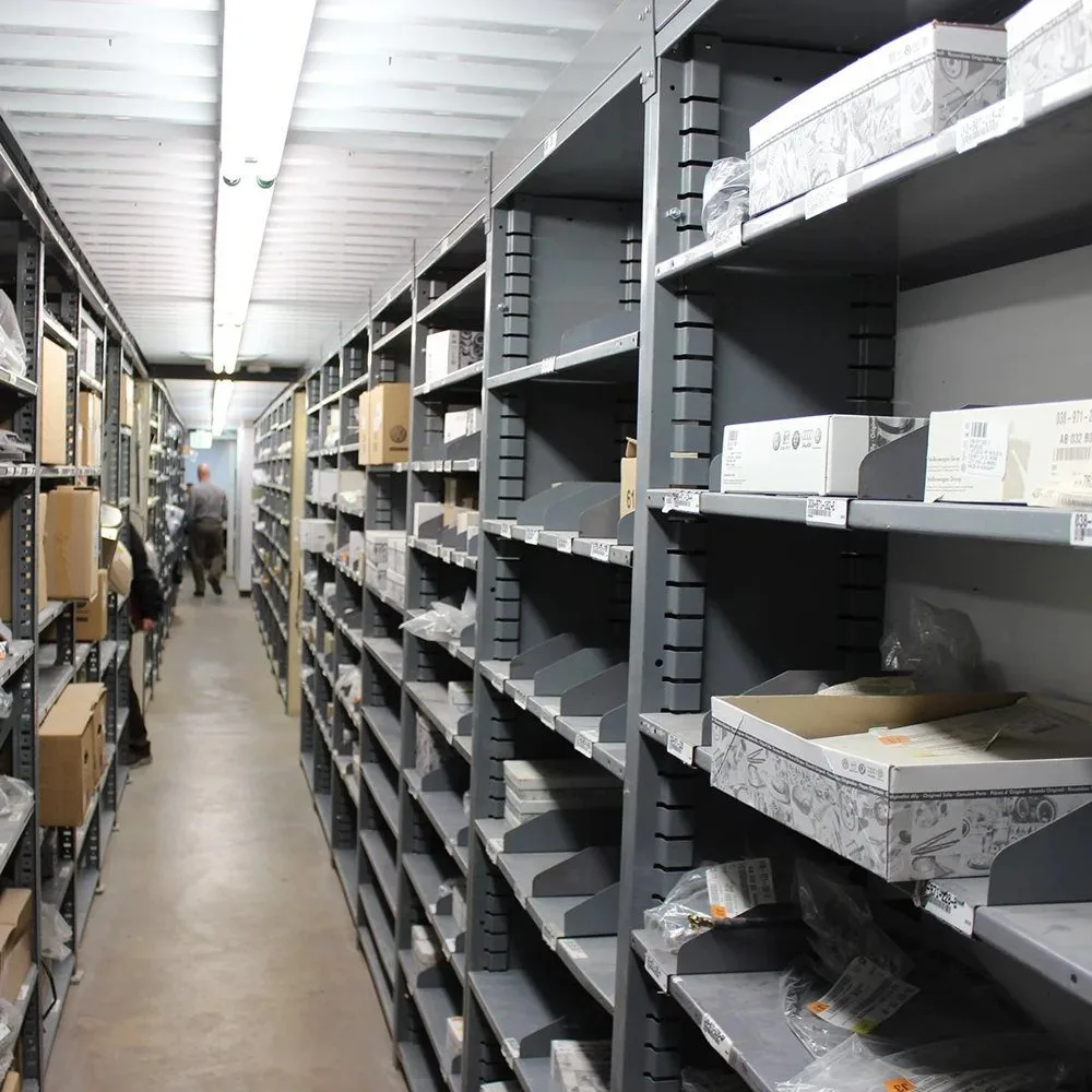 A steel shelving aisle stocked with labeled boxes and parts in a busy stockroom.