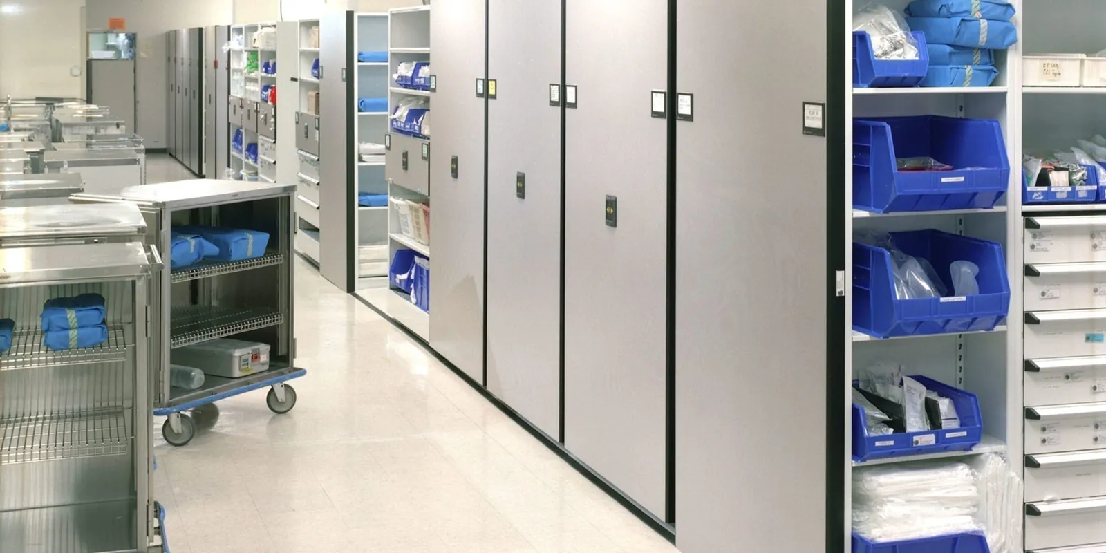 Hospital central supply room with high-density mobile shelving carriages, blue supply bins, and stainless case carts staged in the open floor area
