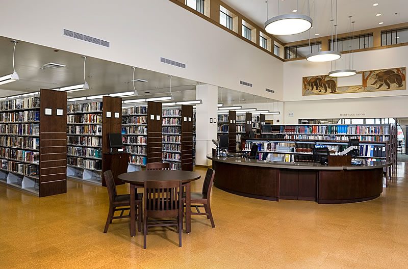 Rows of steel library book storage shelving with wood end panels beside the service desk in a public library, Estey Shelving installation