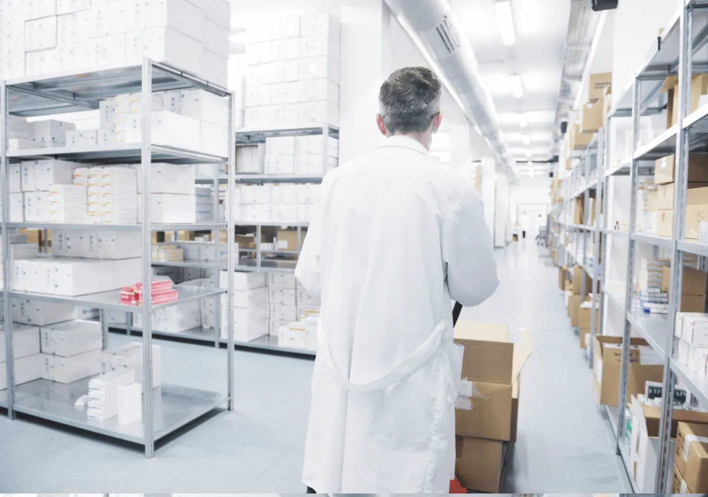 Staff member carrying a carton into a medical supply storage room lined with steel shelving