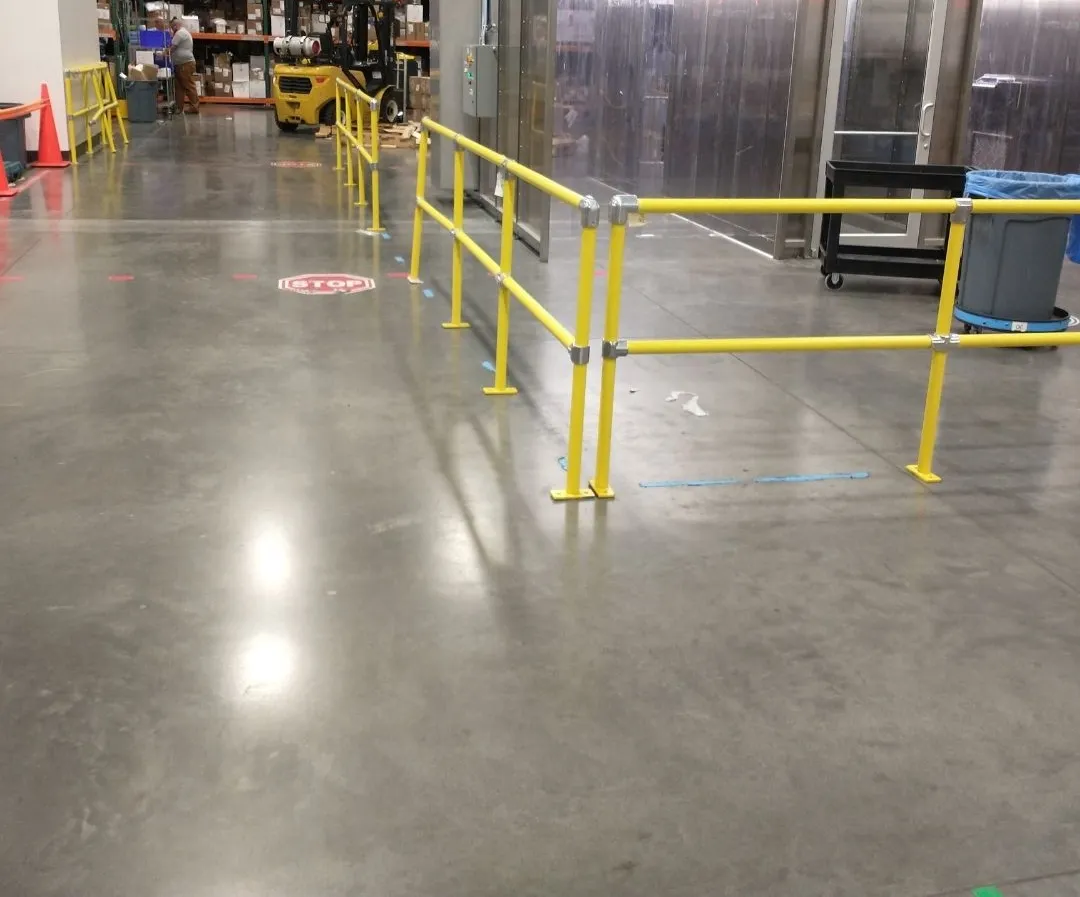Yellow pipe handrail defining a pedestrian walkway beside a forklift aisle, with a stop marking on the floor