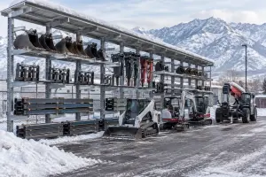 Equipment rental yard in Utah in winter with a covered galvanized cantilever rack holding excavator buckets, forks and shoring rails while loaders and a telehandler park on the plowed lot