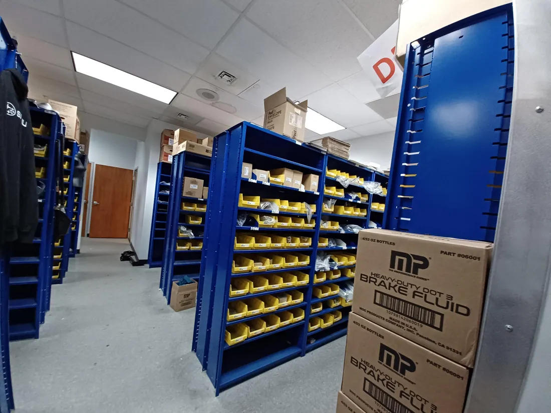 Aisle of existing blue Flexi-Bin units with yellow shelf bins and boxed Subaru parts before the new parts department