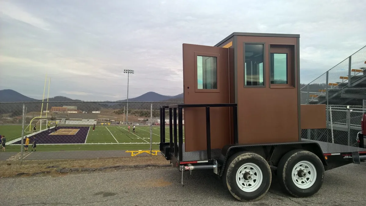 Brown trailer-mounted booth with windows parked beside a school athletic field
