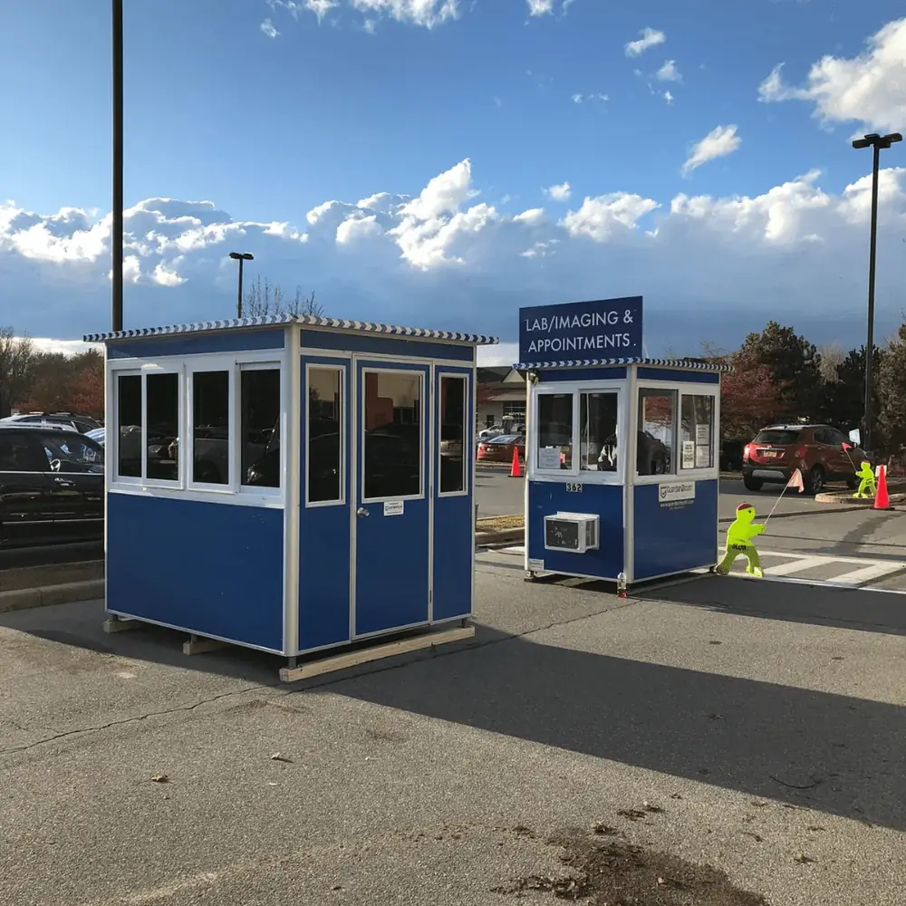 Two blue prefabricated booths with windows set up in a parking lot as a screening station