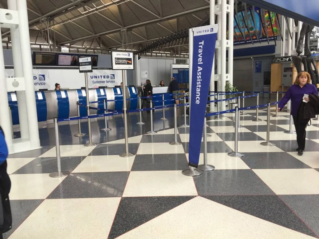 Airport check-in hall with a tall travel assistance sign tower beside stanchion queue lanes