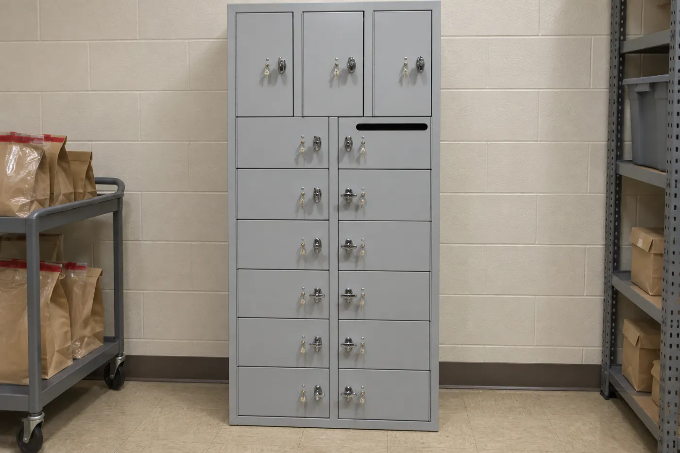 Light gray 15-compartment non-pass-through evidence locker standing against a block wall in a government supply room, beside a cart of sealed evidence bags