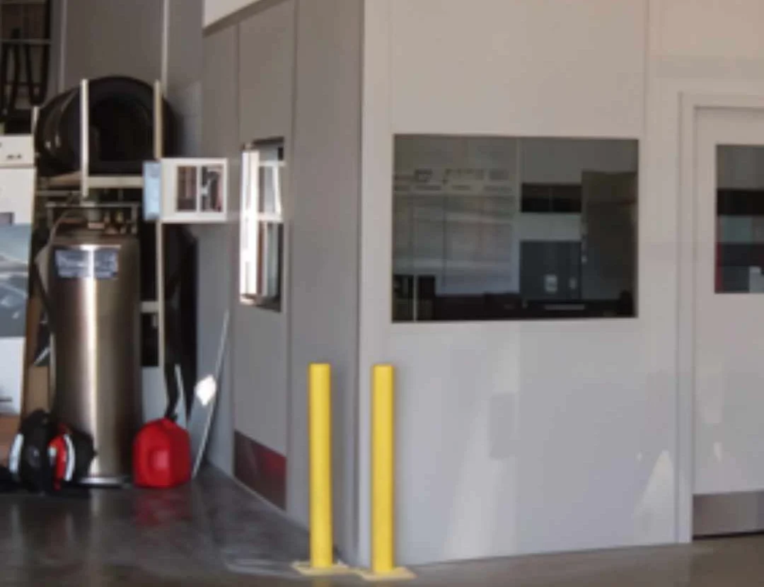 Compact white modular office with a window, protected by yellow bollards inside a plant