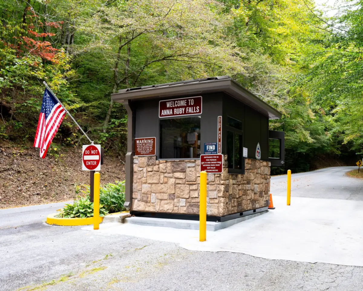 Prefabricated entrance booth with a stone-textured base and dark panel walls beside a paved road at a forested recreation area, protected by yellow bollards