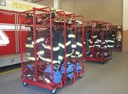 Mobile red turnout gear racks loaded with gear parked beside a fire engine in the apparatus bay