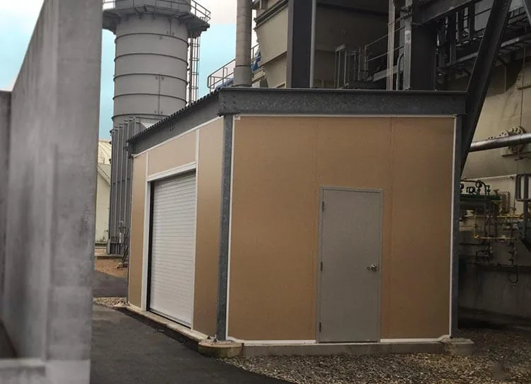 Tan insulated-panel storage building with a roll-up door and a steel personnel door on a curb at a power plant