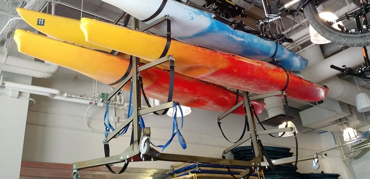 Rec center kayaks strapped to a ceiling-mounted overhead lift frame raised above an equipment room floor