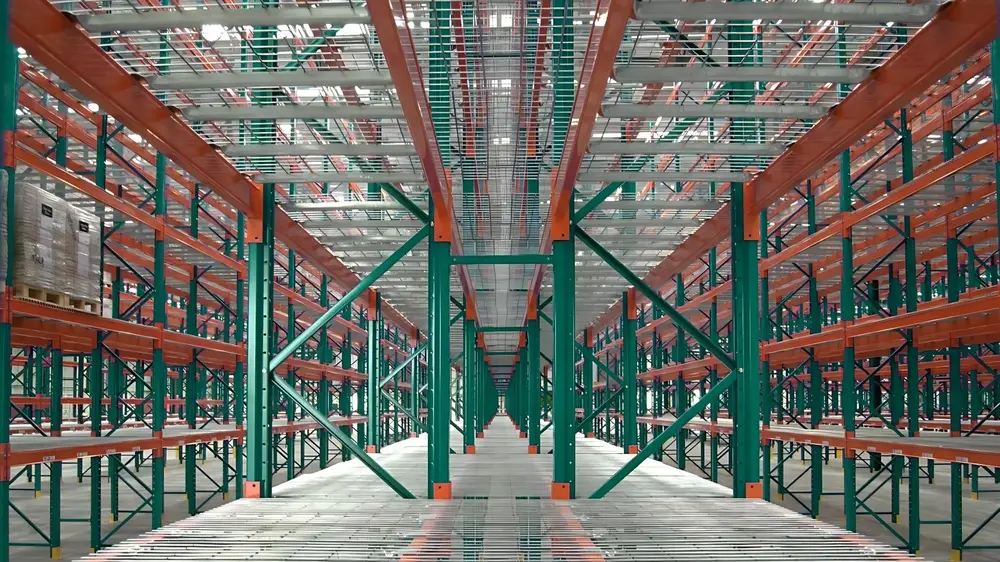 Pallet rack aisle seen from floor level with wire mesh decks on every beam level, showing the open mesh overhead