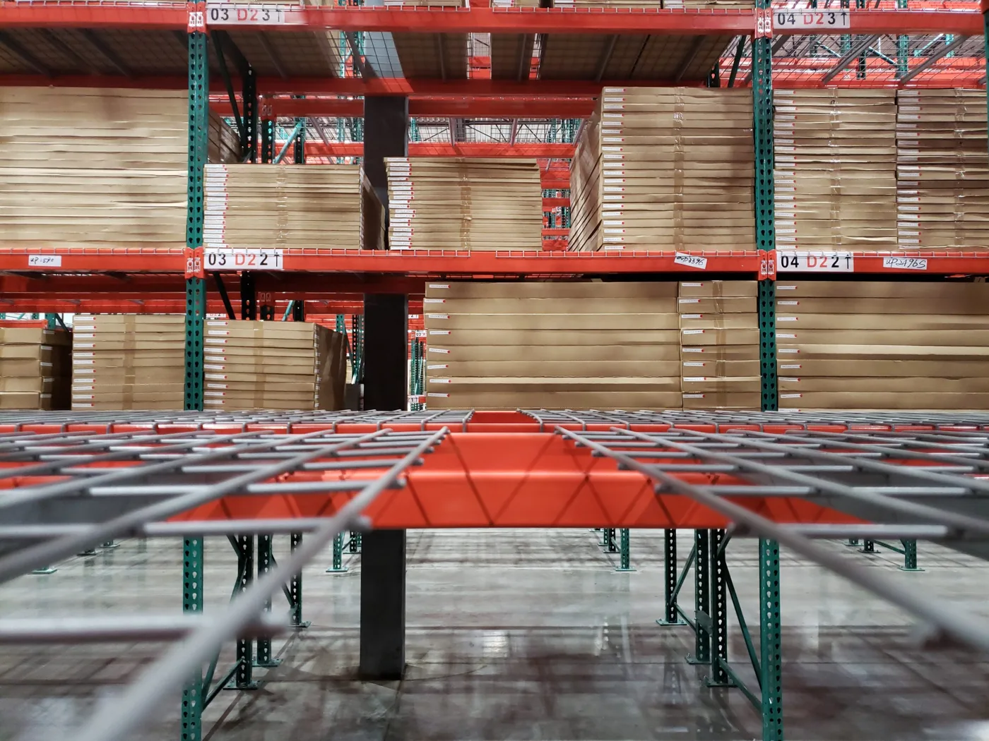 Low view across a wire deck on an orange pallet rack beam, with boxed loads on the levels above and open gaps between loads in each bay